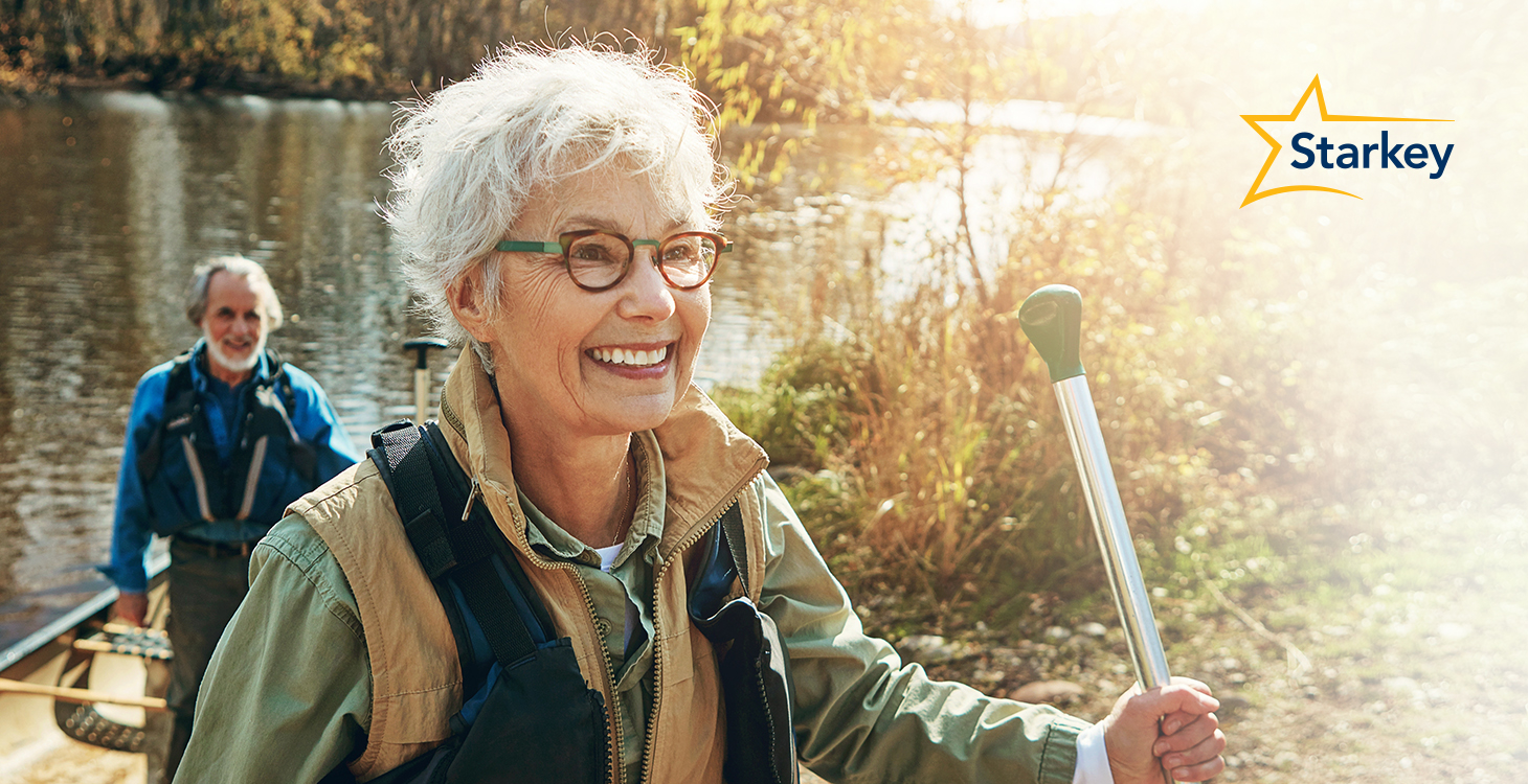 Image of senior woman walking outside with a walking stick with her husband following behind