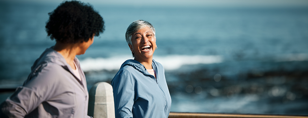 Image of senior woman laughing with younger woman, with ocean waves in the background 